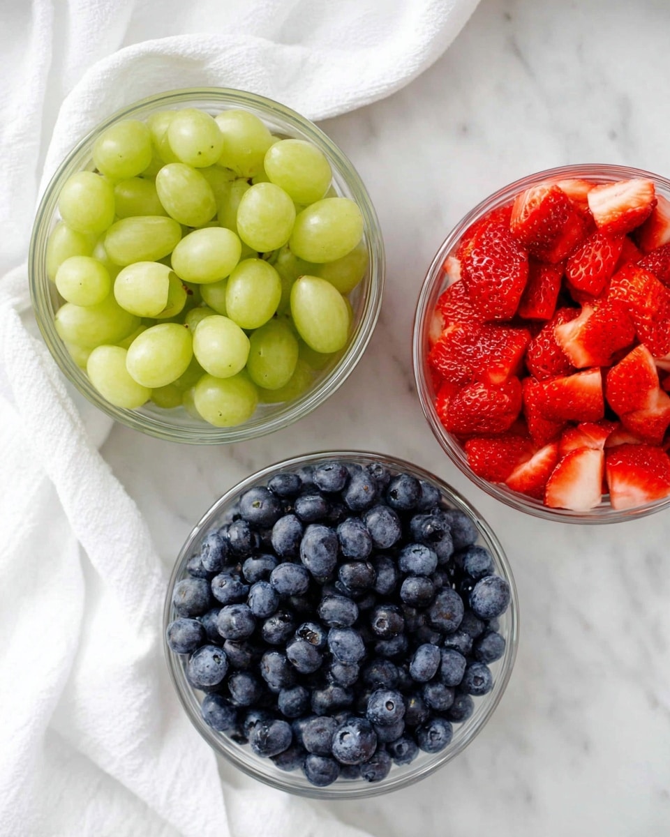 The image shows three clear glass bowls on a white marbled surface; each bowl is filled with different fruit. The top bowl contains light green grapes, smooth and round, filling the bowl fully. The middle bowl holds bright red strawberries, sliced in halves and quarters, showing their juicy texture and seeds visible on the surface. The bottom bowl is packed with plump blue blueberries, displaying their round shape and slightly dusty blue skin. A white cloth is casually placed to the left side of the bowls, adding softness to the scene. photo taken with an iphone --ar 4:5 --v 7