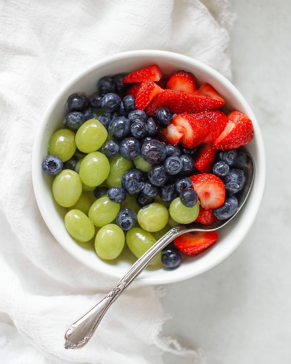 A white bowl filled with three layers of fruit: the bottom layer is dark blue blueberries, the middle layer is large, smooth, light green grapes, and the top layer is bright red, shiny, sliced strawberries. A vintage silver spoon holds some grapes, strawberries, and blueberries and rests partly inside the bowl. The bowl sits on a white marbled surface with a soft white cloth nearby. Photo taken with an iphone --ar 4:5 --v 7