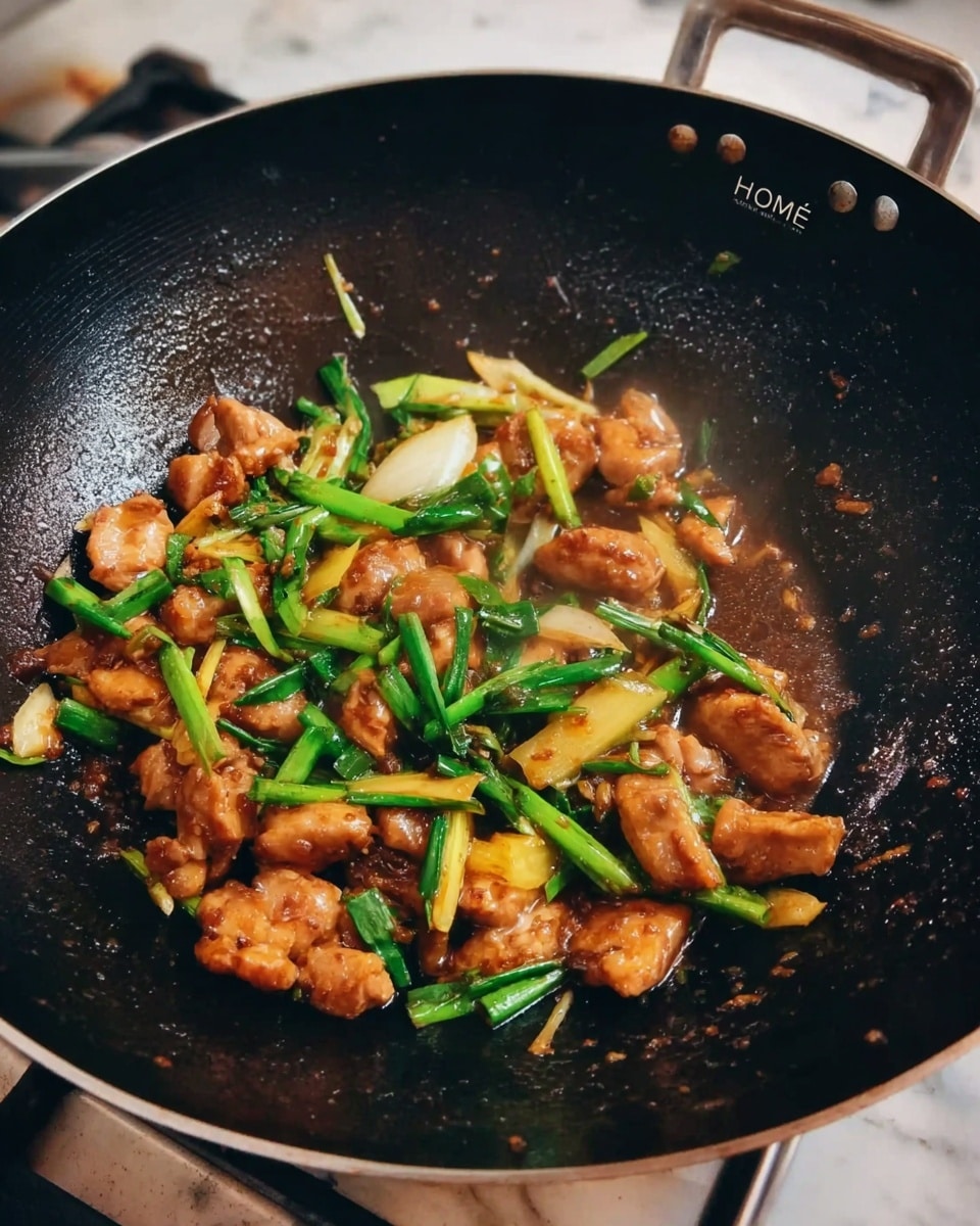 A black wok filled with small pieces of cooked light brown meat mixed with bright green chopped spring onions and slices of light yellow ginger, all coated in a shiny brown sauce. The word