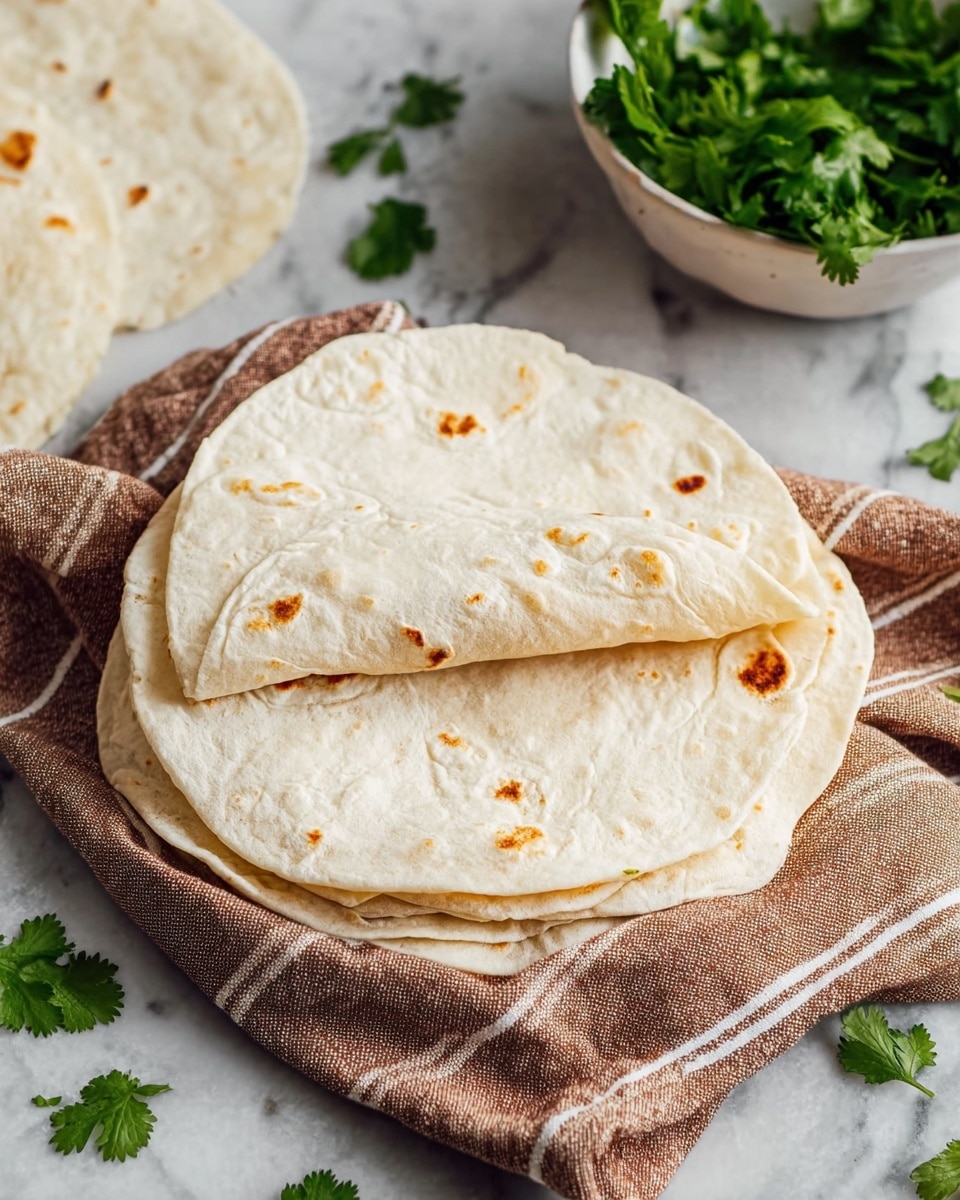The image shows a stack of three round, soft tortillas resting on a brown cloth with white stripes, placed on a white marbled surface. The top tortilla is slightly folded to reveal its thin, smooth texture with small brown toasted spots scattered across its off-white surface. Some fresh green cilantro leaves are scattered around the tortillas and a white bowl filled with more cilantro is visible in the top right corner. The overall scene has a simple and clean look with neutral and natural colors. Photo taken with an iphone --ar 4:5 --v 7
