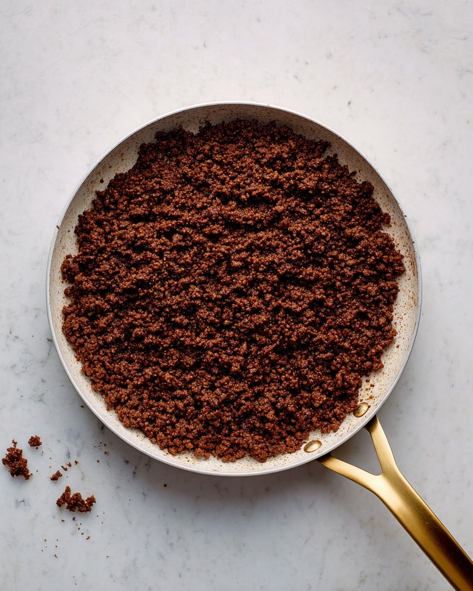 A round white pan with a shiny golden handle is filled with cooked ground meat that is dark brown, slightly crumbly, and spread evenly across the bottom. The pan sits on a white marbled surface with a few small bits of meat scattered near the edges inside the pan, showing a textured and rich finish. The whole scene is viewed from above, highlighting the uniform layer of cooked meat in the pan photo taken with an iphone --ar 4:5 --v 7