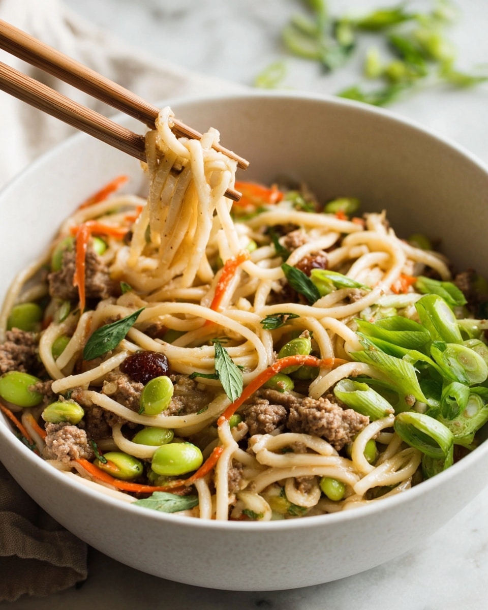 A white bowl filled with a colorful noodle dish, showing thick light beige noodles mixed with small brown meat pieces, bright green edamame beans, thin orange carrot strips, and fresh green herb leaves scattered on top. There are also sliced green onions piled on one side of the bowl. Light wooden chopsticks hold a small bundle of noodles in the center, lifting them slightly above the rest of the food. The bowl sits on a white marbled surface with hints of green herbs blurred in the background. photo taken with an iphone --ar 4:5 --v 7