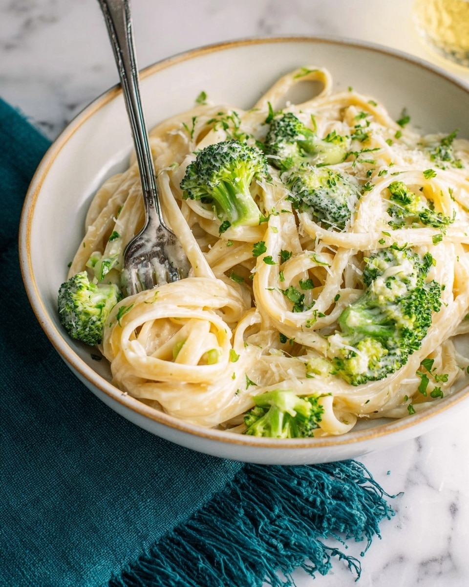 A white shallow bowl holds a serving of creamy pasta with bright green broccoli pieces mixed evenly throughout. The pasta strands are thick and coated in a smooth, glossy white sauce, with small bits of fresh green herbs sprinkled on top. A silver fork is placed in the bowl, partially twirling the pasta. The bowl sits on a dark teal fringed cloth against a white marbled textured surface. Photo taken with an iphone --ar 4:5 --v 7