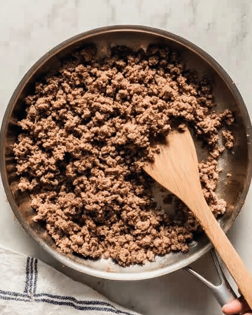 The image shows a silver pan filled with cooked ground meat that is brown and crumbly in texture. A woman's hand holds a wooden spatula inside the pan, positioned on the right side, slightly lifting some of the meat. The pan is placed on a white marbled surface with a small part of a white and black striped cloth visible at the bottom right corner. Photo taken with an iphone --ar 4:5 --v 7
