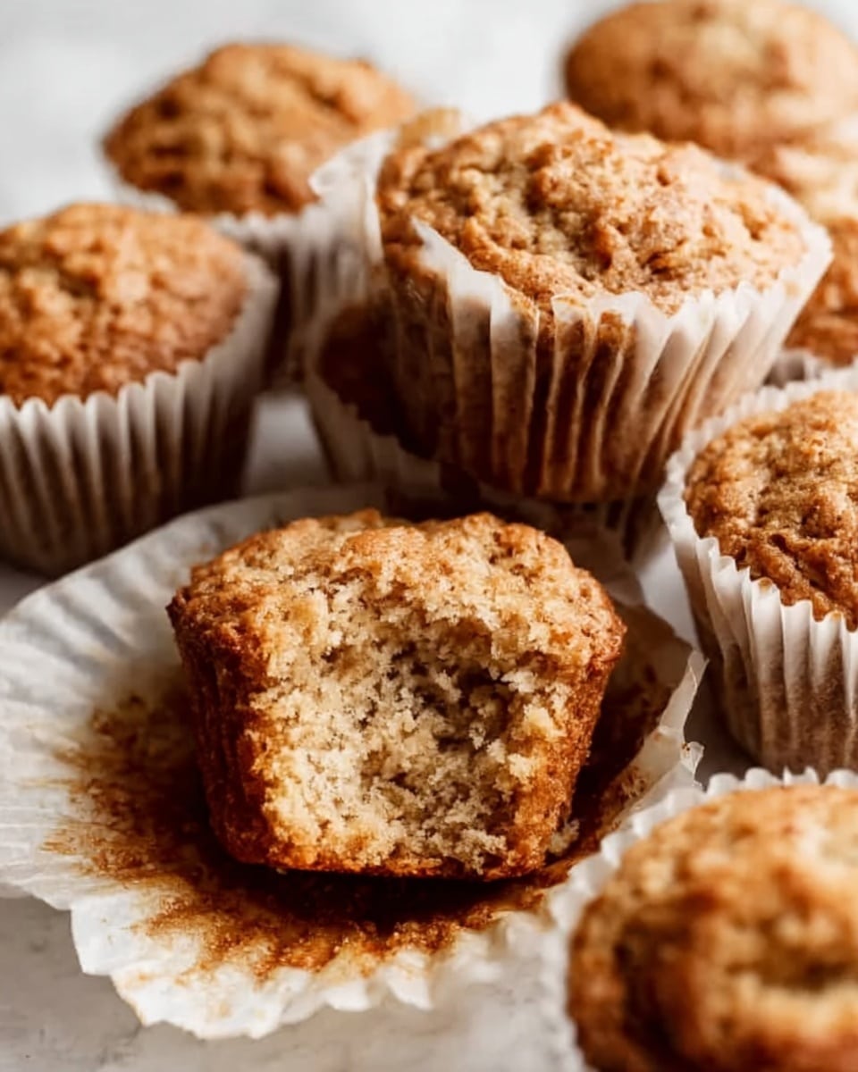 The image shows several muffins in white paper liners arranged closely together on a white marbled surface. One muffin is positioned at the front, partially unwrapped with its liner pulled down and a bite taken out, revealing a soft, crumbly texture inside in light brown shades with darker specks. The undamaged muffins behind it are rough on top with a golden-brown color, looking slightly cracked and rustic. The lighting brings out the warm tones and texture details of the muffins, emphasizing their tender crumb and slightly coarse surface. Photo taken with an iphone --ar 4:5 --v 7