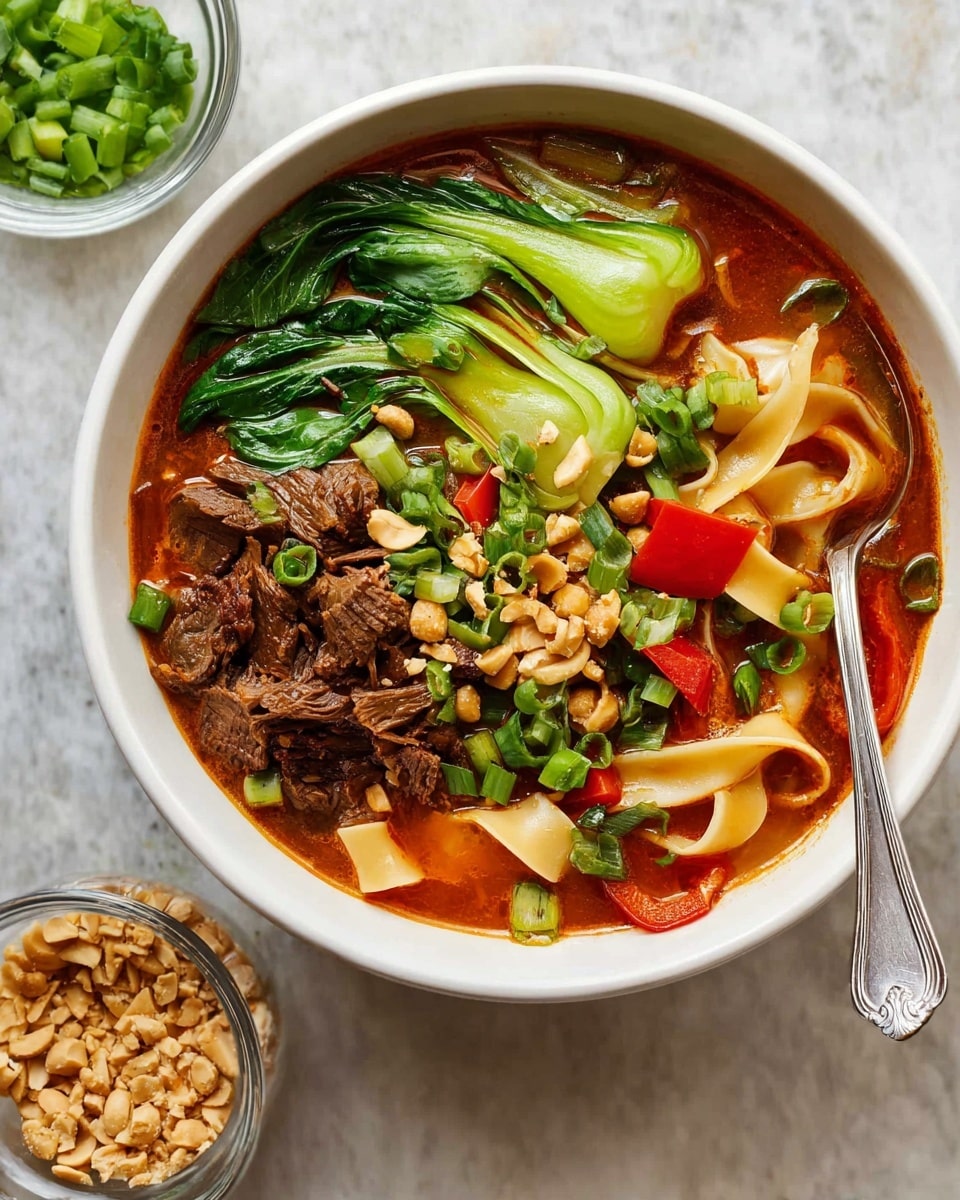A white bowl filled with a rich reddish-orange broth holds wide flat noodles curled on the left side, with green bok choy leaves and bright red bell pepper slices floating near the top. Tender shredded dark brown meat pieces are placed mostly on the right side, mixed with chopped green onions scattered across the top. Small crushed peanuts are sprinkled over the dish. A small white bowl with chopped green onions sits above the main bowl, and a glass bowl with more crushed peanuts is placed below it, all set on a white marbled surface. A spoon rests inside the bowl. photo taken with an iphone --ar 4:5 --v 7