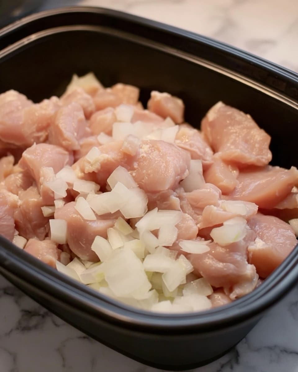 The image shows raw diced chicken pieces mixed with small, chopped white onion pieces inside a black container. The chicken pieces are light pink, soft-looking, and scattered evenly with the firm, slightly translucent white onion bits. The container has a smooth, matte texture and rounded edges. The background is a white marbled surface. photo taken with an iphone --ar 4:5 --v 7