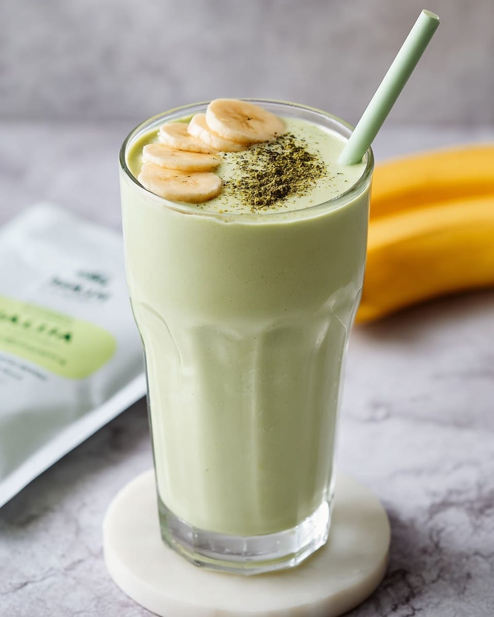 A tall clear glass filled with a light green smoothie, smooth and creamy in texture, topped with three round pale yellow banana slices arranged near the center. On top of the banana slices, there is a small sprinkle of dark green powder. A pale green straw is inserted into the drink from the right side. The glass sits on a white round coaster, placed on a white marbled surface. A yellow banana and a white package with green text are blurred in the soft background. Photo taken with an iphone --ar 4:5 --v 7