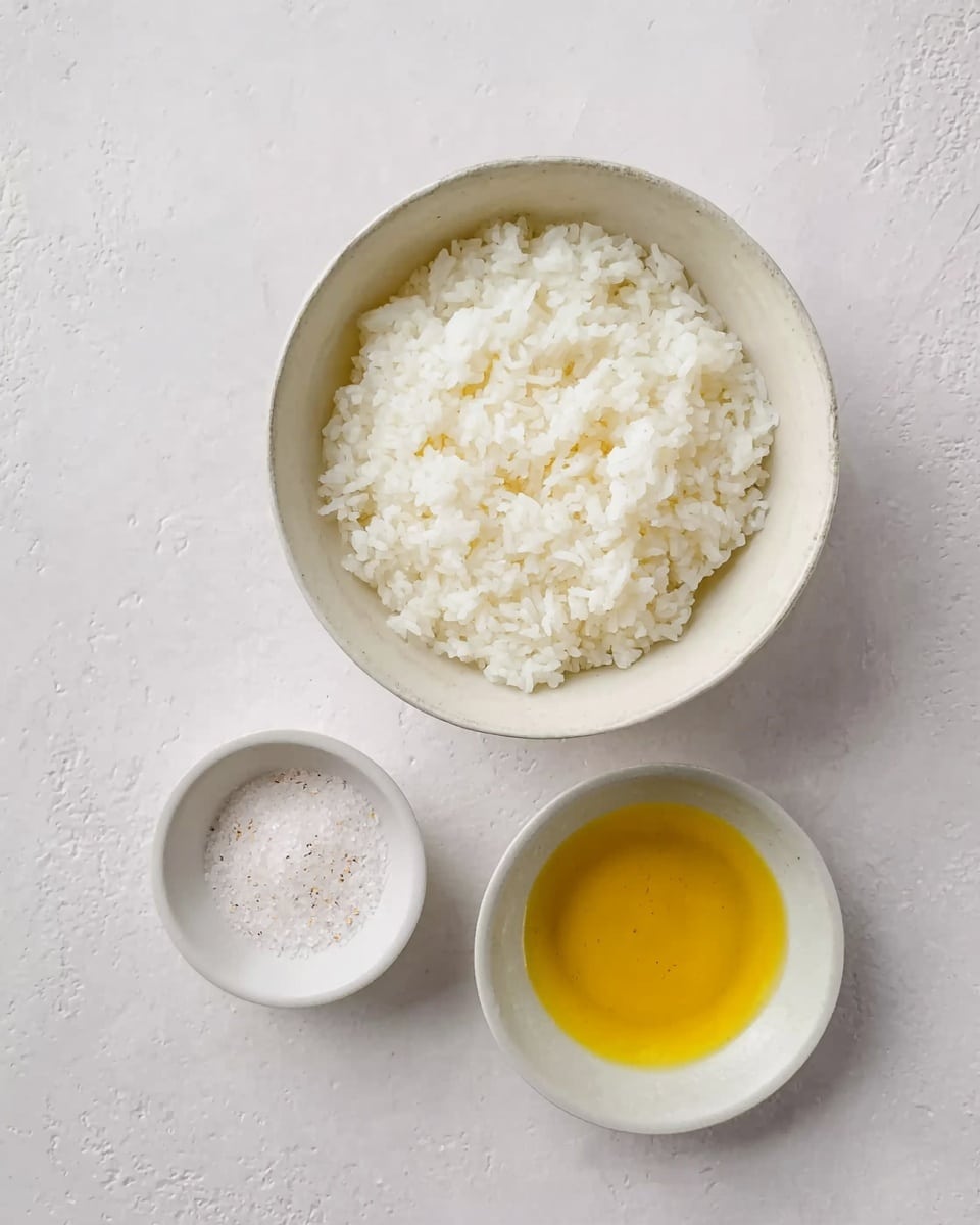 The image shows a white bowl filled with cooked white rice placed on a white marbled surface. Below the bowl, there are two small white bowls; the one on the left contains a white granular substance, likely salt, and the one on the right holds a golden-yellow liquid, most likely oil. The overall look is clean and simple with soft natural light. photo taken with an iphone --ar 4:5 --v 7