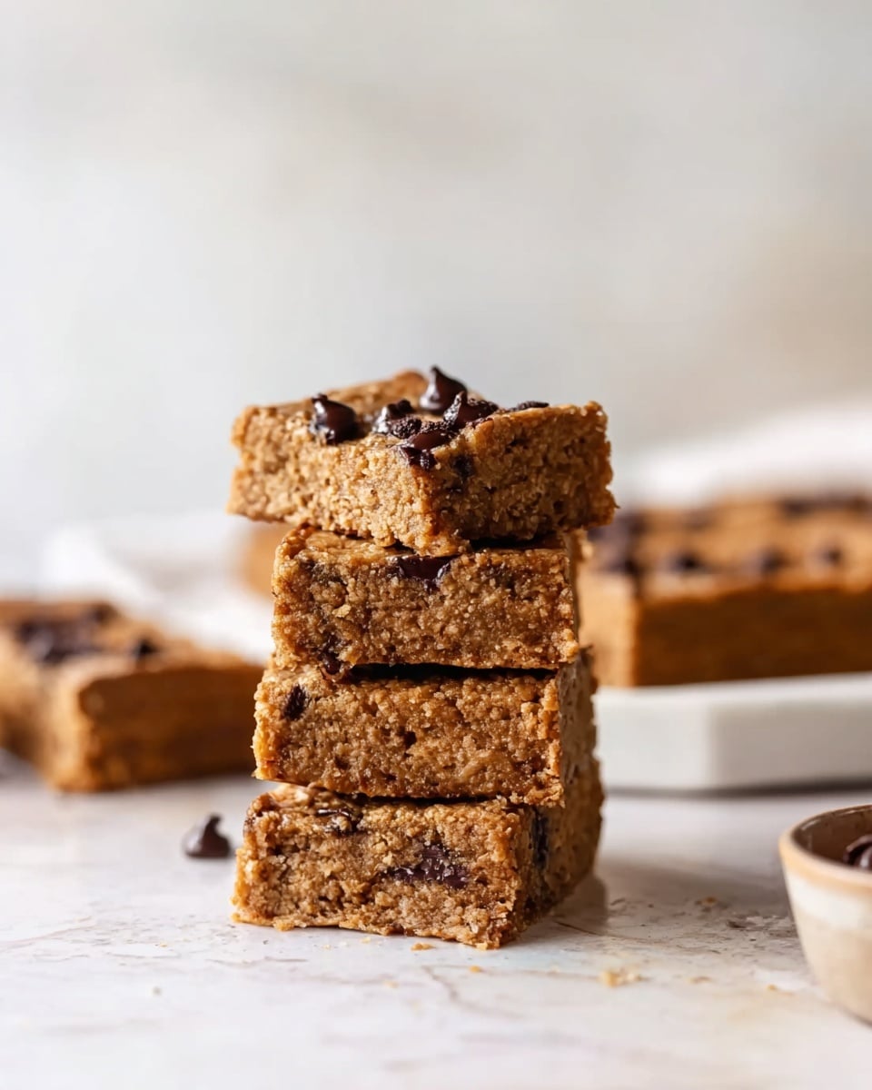 The image shows a stack of four thick, square bars with a golden brown color and visible chocolate chips inside and on top, creating a slightly glossy texture. Each bar has a chewy, soft look with a slightly crumbly edge, and the top bar is slightly tilted. In the background, there are more bars on a white marbled surface with a blurred white wall behind. The lighting is soft and natural, giving the bars a warm and inviting appearance. Photo taken with an iphone --ar 4:5 --v 7