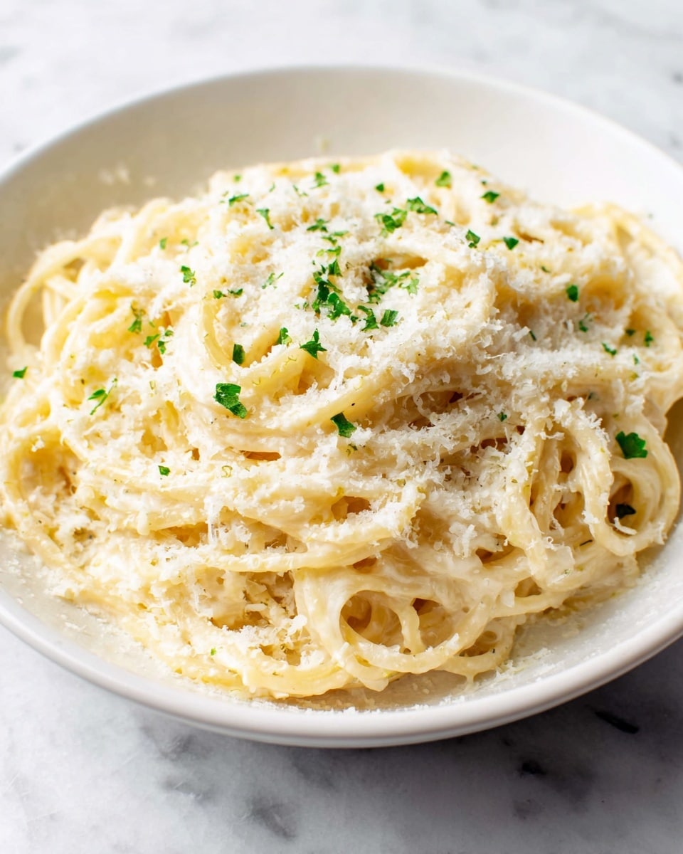 A close-up of a white bowl filled with creamy spaghetti pasta, coated evenly with a smooth light cream sauce. The top layer shows a generous amount of finely grated white cheese sprinkled all over the pasta, with small green herb flakes scattered lightly for color. The pasta strands are thick and curled naturally, creating a soft textured look beneath the cheese. The bowl sits on a white marbled surface, making the whole dish look fresh and simple. photo taken with an iphone --ar 4:5 --v 7