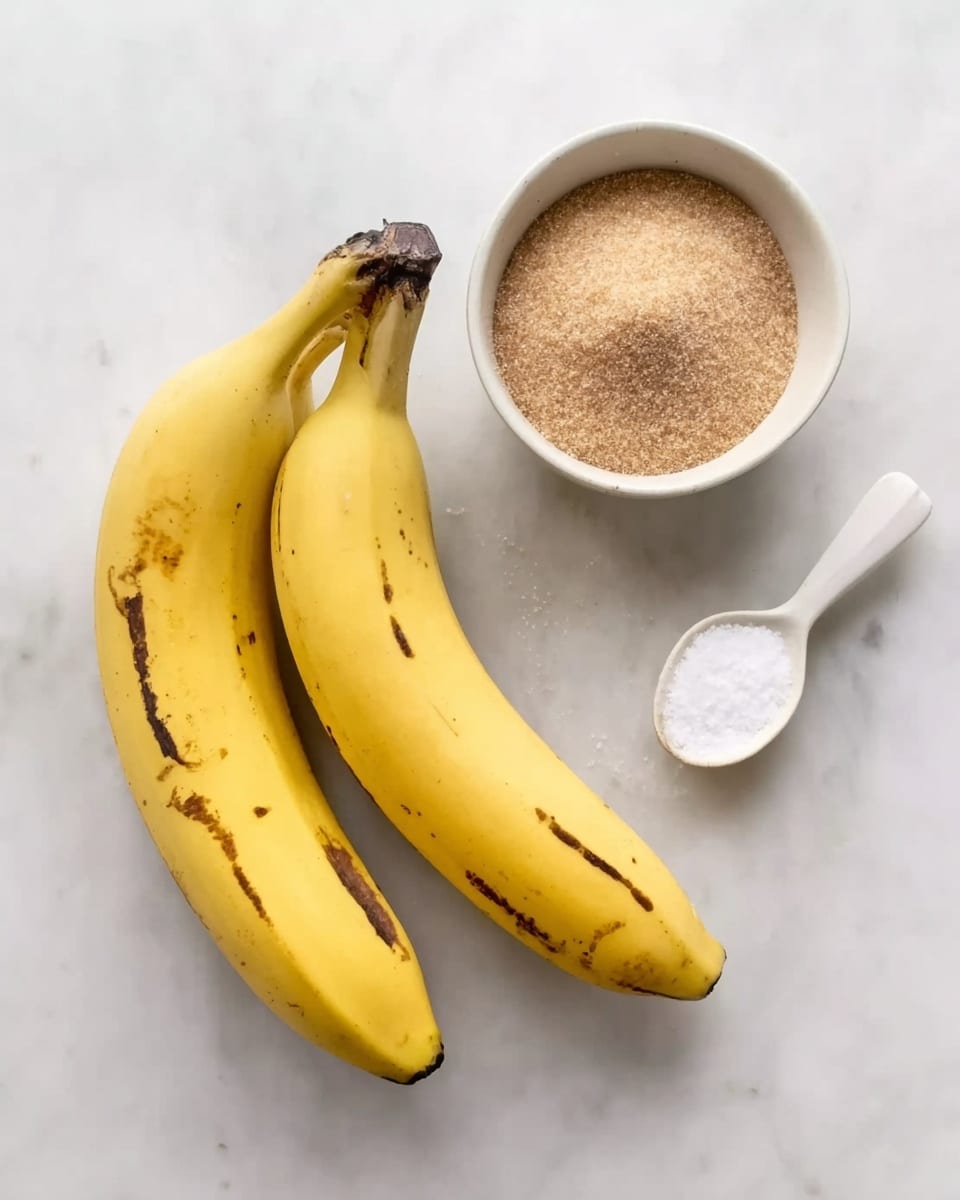 The image shows two ripe yellow bananas with some brown spots, placed side by side on a white marbled surface. To the left of the bananas is a white bowl filled with brown sugar, and to the right is a small white bowl containing salt. A white spoon with its handle pointing to the bottom right corner rests between the bananas and the salt bowl. The overall setup is simple, clean, and bright, with a neutral color palette. photo taken with an iphone --ar 4:5 --v 7