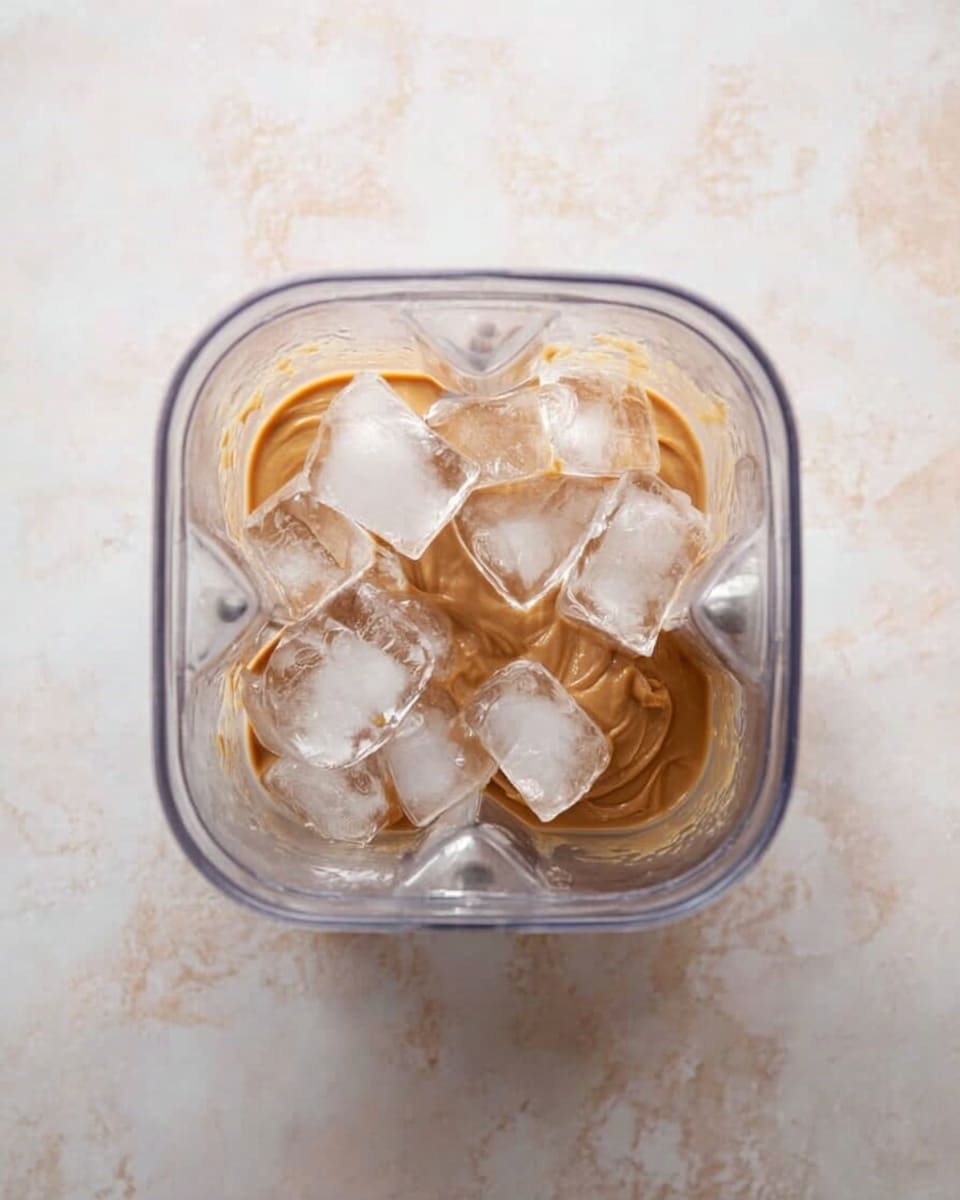 A clear blender container is shown from above, holding a layer of light brown peanut butter-like paste at the bottom, topped with a layer of irregular, glossy, translucent ice cubes. The container sits on a white marbled surface. Photo taken with an iphone --ar 4:5 --v 7