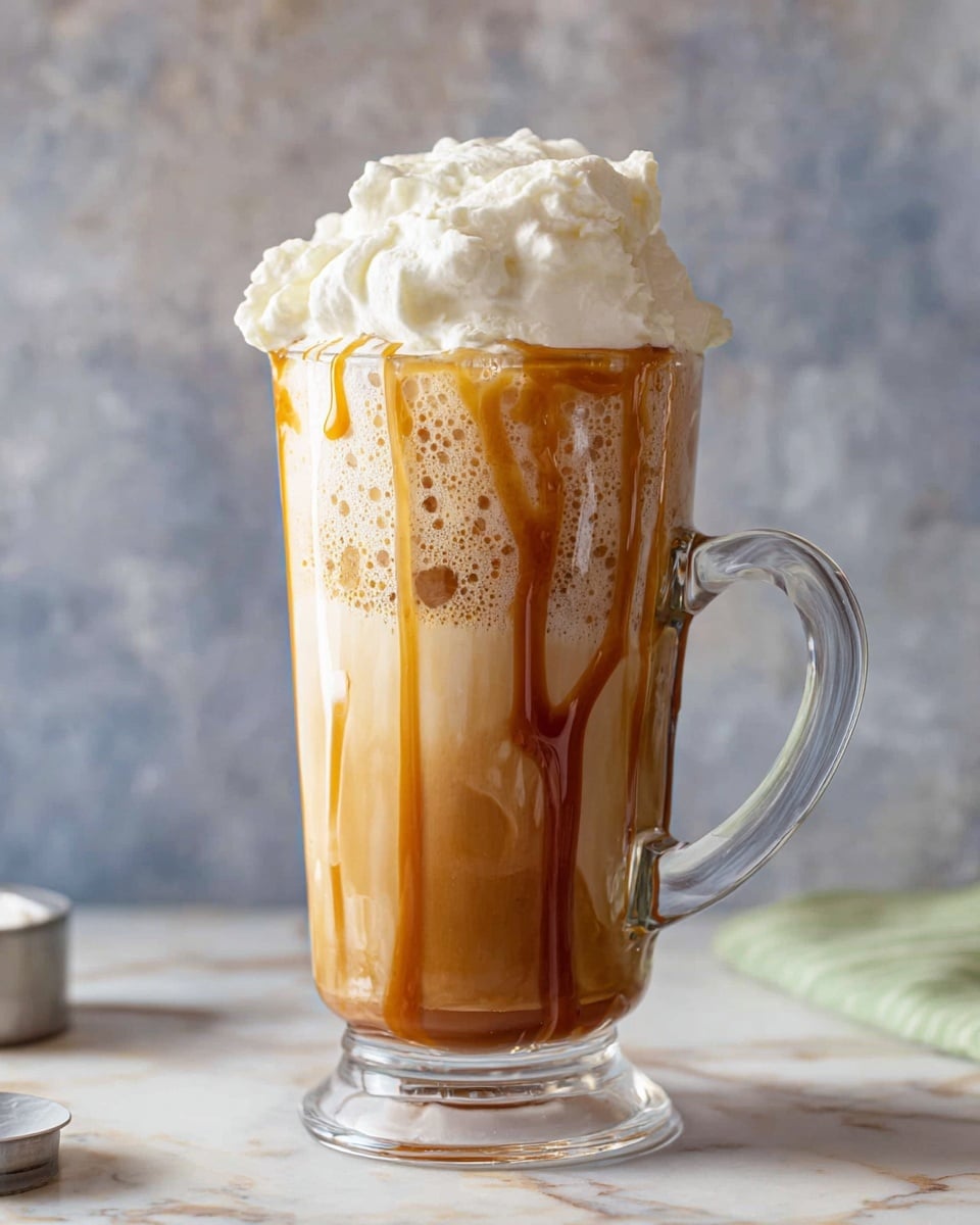 A tall clear glass mug shows a three-layer drink on a white marbled surface. The bottom layer is smooth and light brown with caramel sauce streaks dripping down inside the glass. The middle layer is a frothy, airy mix of light brown and beige, looking bubbly and creamy. The top layer is a thick, fluffy white cream piled high above the glass rim, slightly uneven and soft. The clear handle on the right side adds to the clean look, and the background is a blurred gray textured wall. Photo taken with an iphone --ar 4:5 --v 7