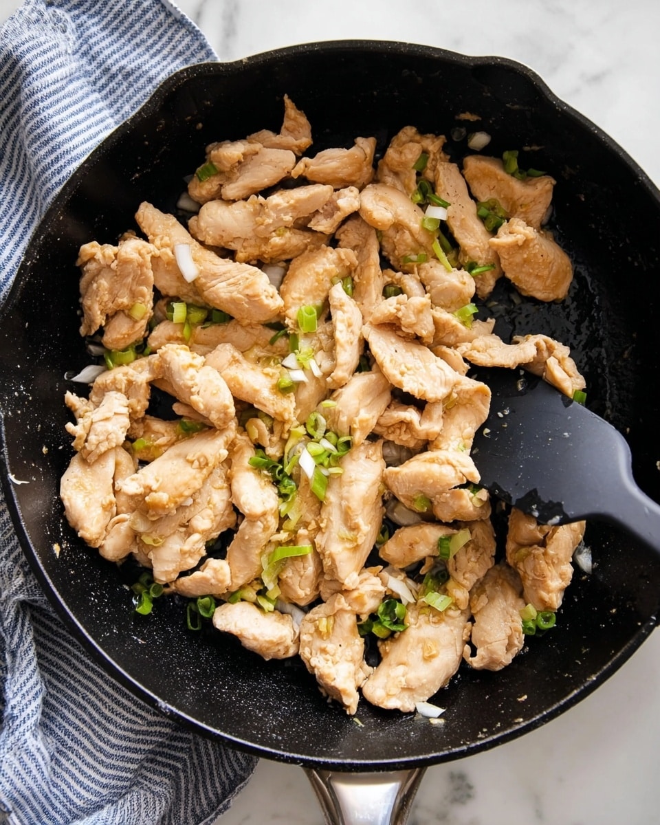 The image shows a black cast iron pan filled with cooked slices of light brown chicken mixed with small pieces of green onions and finely chopped white onions. The chicken pieces have a soft, slightly shiny texture and are piled evenly, covering the pan's surface. A black spatula is lifting some chicken on the right side of the pan, and a blue and white striped cloth is partially visible in the top left corner. The pan sits on a white marbled surface. Photo taken with an iphone --ar 4:5 --v 7