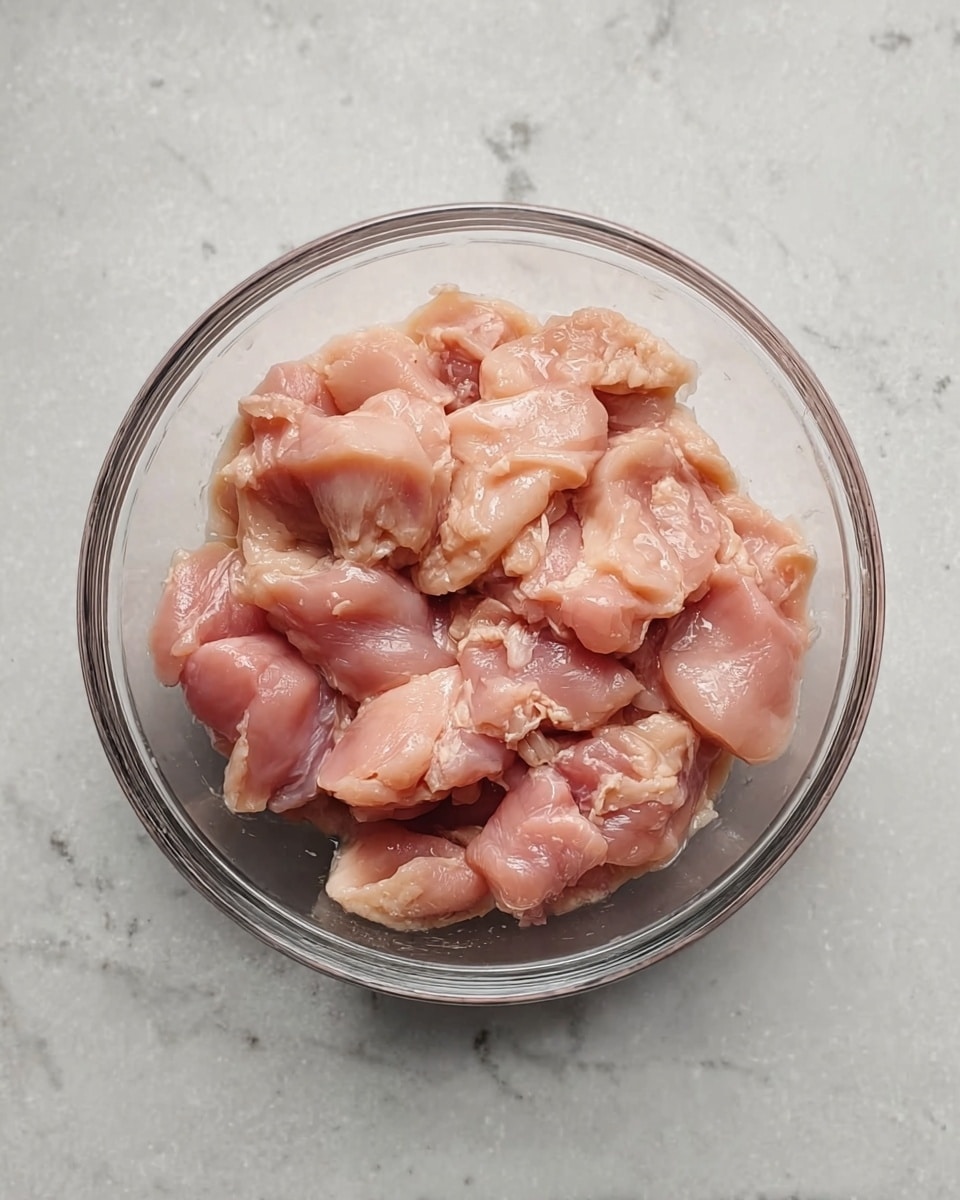 A clear glass bowl is filled with many pieces of raw pale pink chicken with a slight glossy shine, showing different smooth and soft textures. The chicken pieces are piled up inside the bowl, with some overlapping and others lying flat. The bowl sits on a white marbled surface that has faint gray veins, creating a clean and simple background. photo taken with an iphone --ar 4:5 --v 7