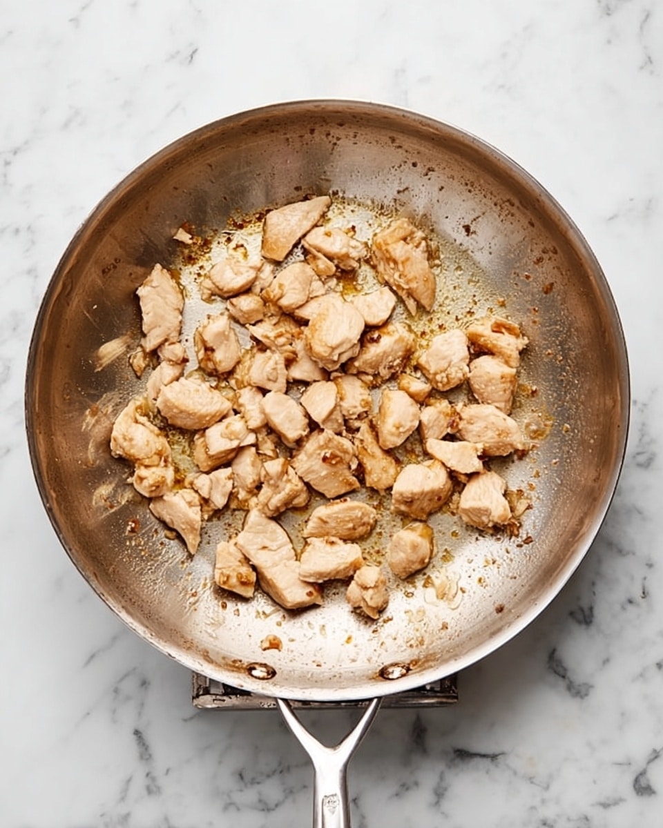 A silver frying pan with many small pieces of light brown cooked chicken scattered unevenly inside, showing some browned spots and a few golden bits stuck to the pan's surface. The pan has two metal handles, and it sits on a white marbled tabletop. The chicken pieces have a slightly rough texture and are not stacked but spread out in a single layer. photo taken with an iphone --ar 4:5 --v 7