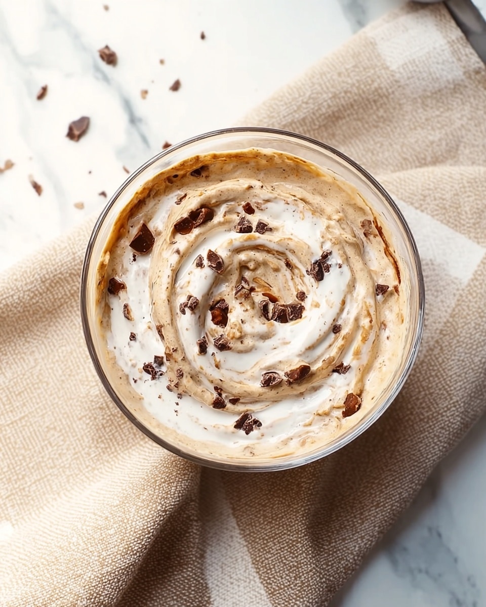 A clear glass bowl holds a creamy mix with two visible layers swirled together: a light beige base and a white creamy layer mixed in a spiral pattern on top. Small dark brown pieces, likely chocolate or nuts, are scattered across the surface, adding texture. The bowl is placed on a beige towel with a checkered pattern, and the background shows a white marbled surface with a few scattered dark crumbs. Photo taken with an iphone --ar 4:5 --v 7