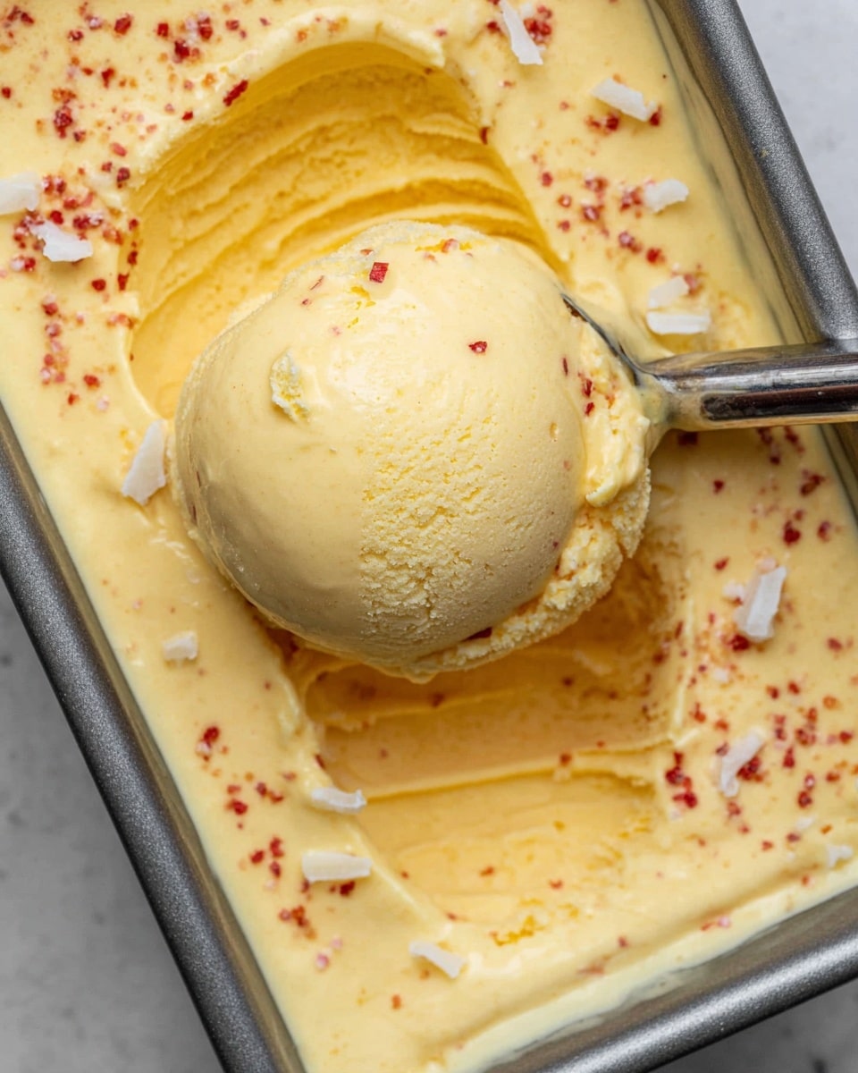A close-up view of a metal baking pan filled with smooth, creamy yellow ice cream with a soft texture. The ice cream is sprinkled with small red specks and a few white flakes on top. A spoon holds a rounded scoop of the ice cream, positioned above the pan's surface, showing the rich and thick consistency. The background is a white marbled texture. photo taken with an iphone --ar 4:5 --v 7