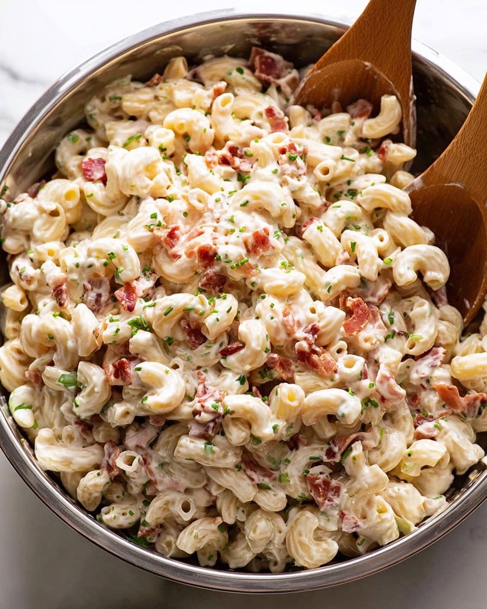 The image shows a large silver metal bowl filled with creamy macaroni salad. The salad has elbow-shaped pasta coated in a thick white sauce, mixed with small pieces of red bacon and finely chopped green herbs sprinkled throughout. Two wooden salad spoons with smooth textures are partially inserted into the bowl from the right side, resting on the pasta. The background is a white marbled surface, giving a clean and bright look. Photo taken with an iphone --ar 4:5 --v 7