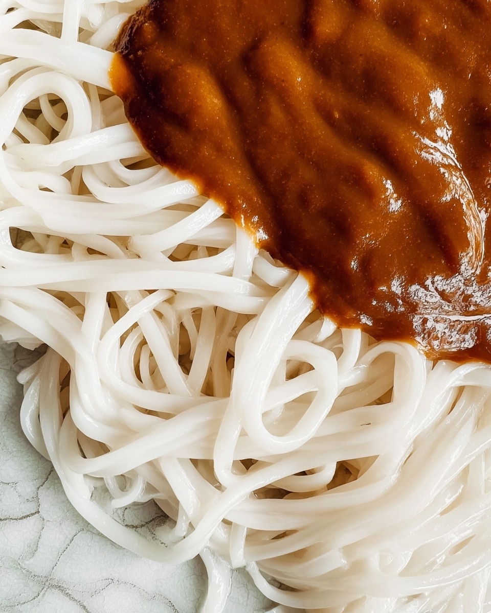This image shows one layer of white noodles with a smooth and slightly shiny texture. On top of the noodles, there is one layer of thick brown sauce that looks creamy and rich with some small darker spots. The noodles are long and flat, folded and intertwined, covering the bottom part of the image. The sauce is placed near the center and spills slightly onto the noodles. The background is a white marbled texture. photo taken with an iphone --ar 4:5 --v 7