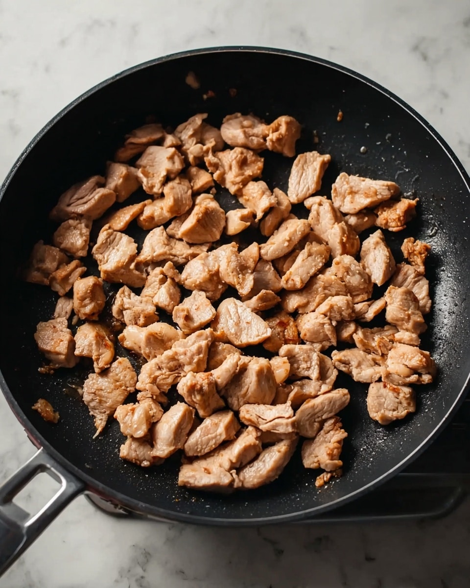 A black frying pan is filled with multiple small pieces of light brown cooked meat spread evenly across its surface. The meat pieces have a soft texture with slightly browned edges, and the pan is set on a white marbled surface. photo taken with an iphone --ar 4:5 --v 7