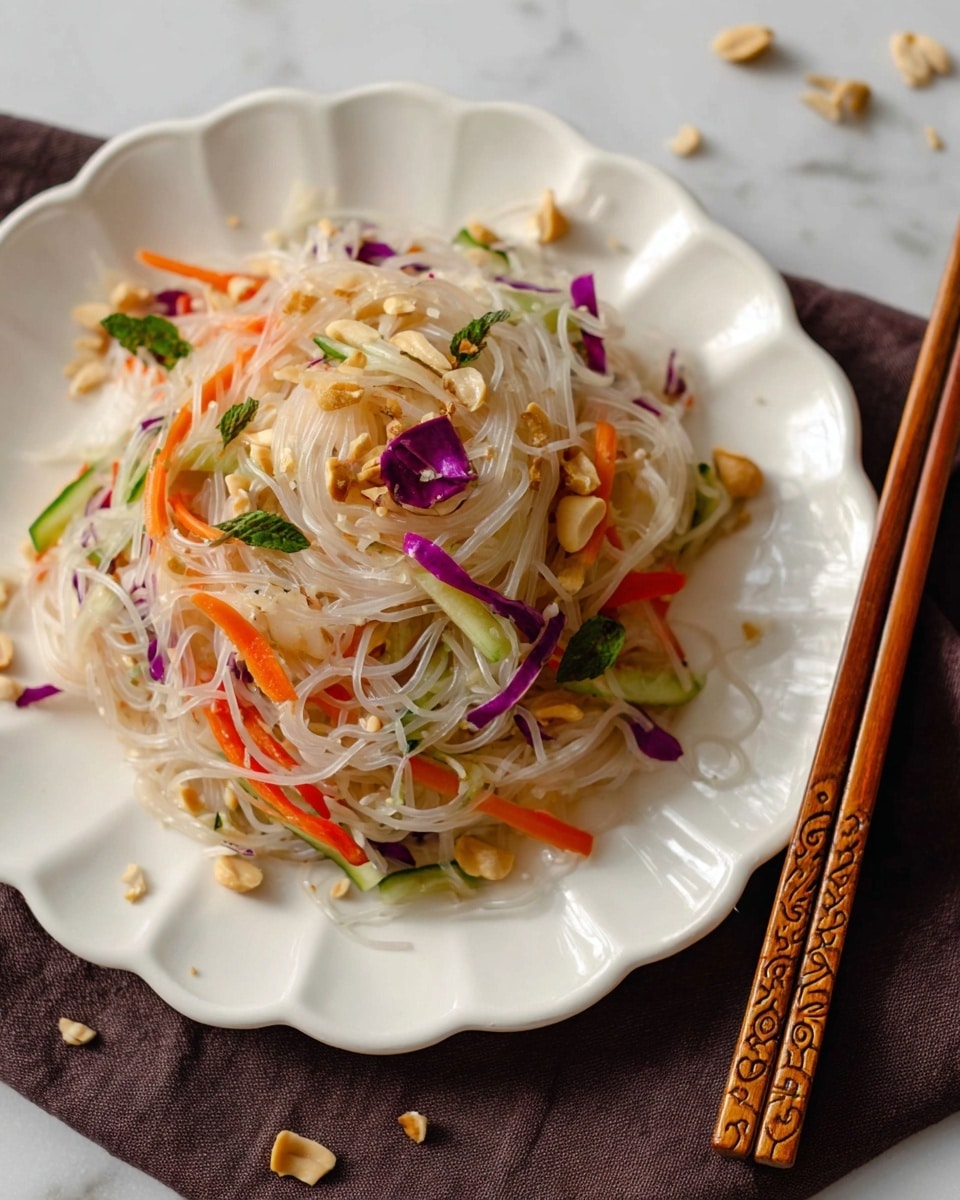 A white plate with a scalloped edge holds a mound of thin, translucent rice noodles mixed with thin strips of orange carrot, green cucumber, and purple cabbage, topped with crushed peanuts and small green herb leaves. The noodles shine slightly, showing a light coating of dressing, and the crunchy peanut pieces are scattered on top and around the plate. A pair of wooden chopsticks with a carved pattern rests near the bottom right edge of the plate, which sits on a dark brown cloth over a white marbled surface. photo taken with an iphone --ar 4:5 --v 7