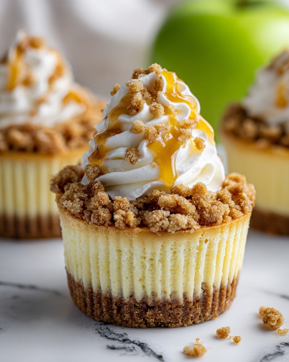 A close-up of a cupcake with three layers on a white marbled surface. The bottom layer is a crumbly, brown crust. The middle layer is a smooth, pale yellow cheesecake filling. The top layer is a rough, crumbly streusel with small nut pieces. On top of the streusel is a dollop of white whipped cream with a drizzle of golden caramel sauce. In the background, there is a green apple that is out of focus, and part of another cupcake. Photo taken with an iphone --ar 4:5 --v 7