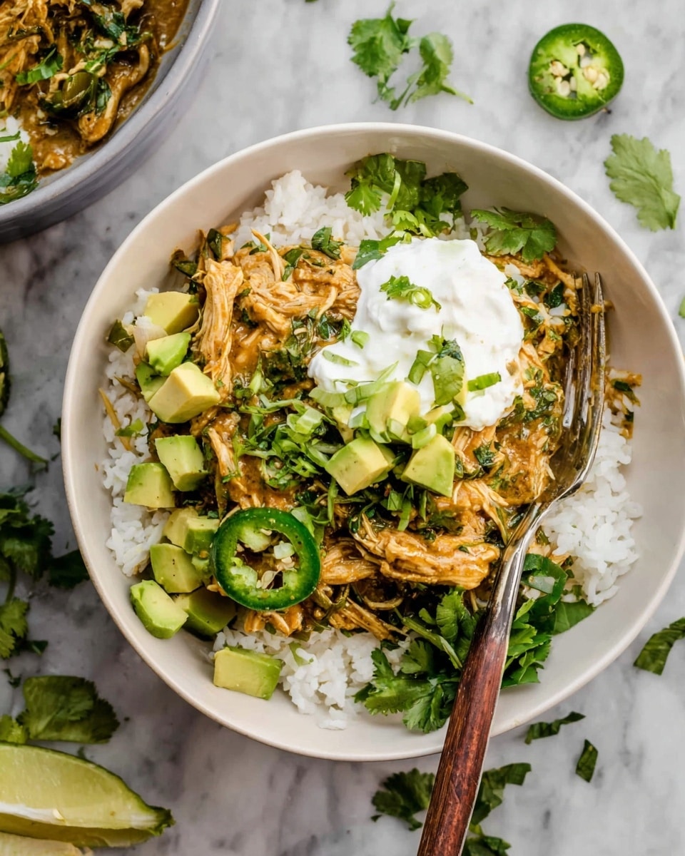 A white bowl filled with a base layer of white rice, topped with shredded chicken mixed with green herbs and a sauce that has an orange-brown color. On top of the chicken, there are several small cubes of avocado and a white dollop of sour cream garnished with small green onion pieces. Around the edges of the bowl, fresh cilantro leaves and sliced green jalapeños are scattered. A fork with a wooden handle is placed inside the bowl. The bowl is on a white marbled surface with another bowl partially visible nearby. Photo taken with an iphone --ar 4:5 --v 7