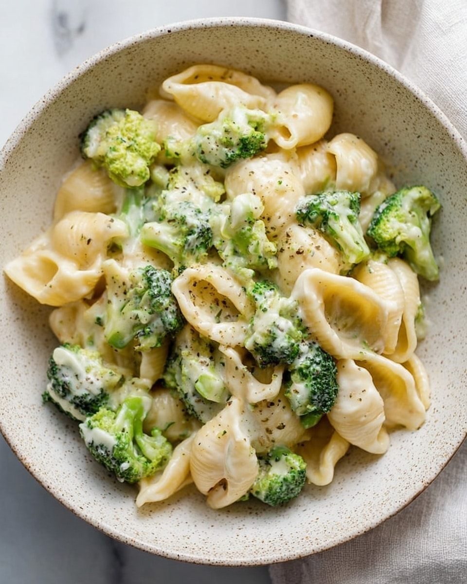 The dish is a close-up view of a white speckled bowl filled with pasta shells mixed with broccoli florets. The pasta shells are light yellow, smooth, and coated in a creamy white sauce that also covers the bright green broccoli pieces, creating a soft and creamy texture throughout. The broccoli adds a fresh pop of green with small, textured buds and firm stems. The bowl sits on a white marbled surface, and in the top corner, part of a light cloth is softly visible. photo taken with an iphone --ar 4:5 --v 7
