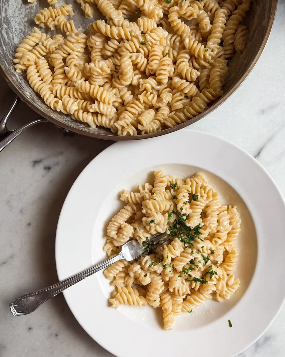 The image shows a simple plate of pasta made with small spiral-shaped noodles covered in a light cream sauce. The pasta is neatly arranged in a white plate with some small green herbs sprinkled on top, adding a touch of color. Next to the plate is a pan containing more pasta of the same kind, coated in the same sauce. A silver fork rests inside the pasta on the plate. All items are placed on a white marbled surface. Photo taken with an iphone --ar 4:5 --v 7