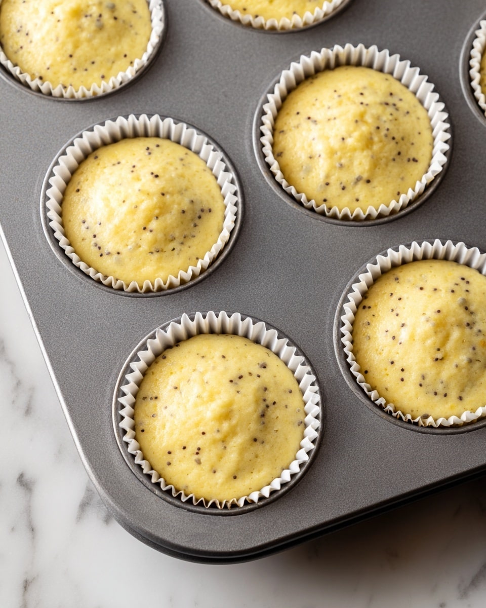 A gray metal muffin tray filled with six round white paper liners, each liner half full of light yellow batter speckled with small dark seeds, sitting on a white marbled surface. The batter looks smooth with a few small lumps in every liner, and the muffin tray is positioned diagonally with the bottom right liner slightly cut off at the edge of the image photo taken with an iphone --ar 4:5 --v 7