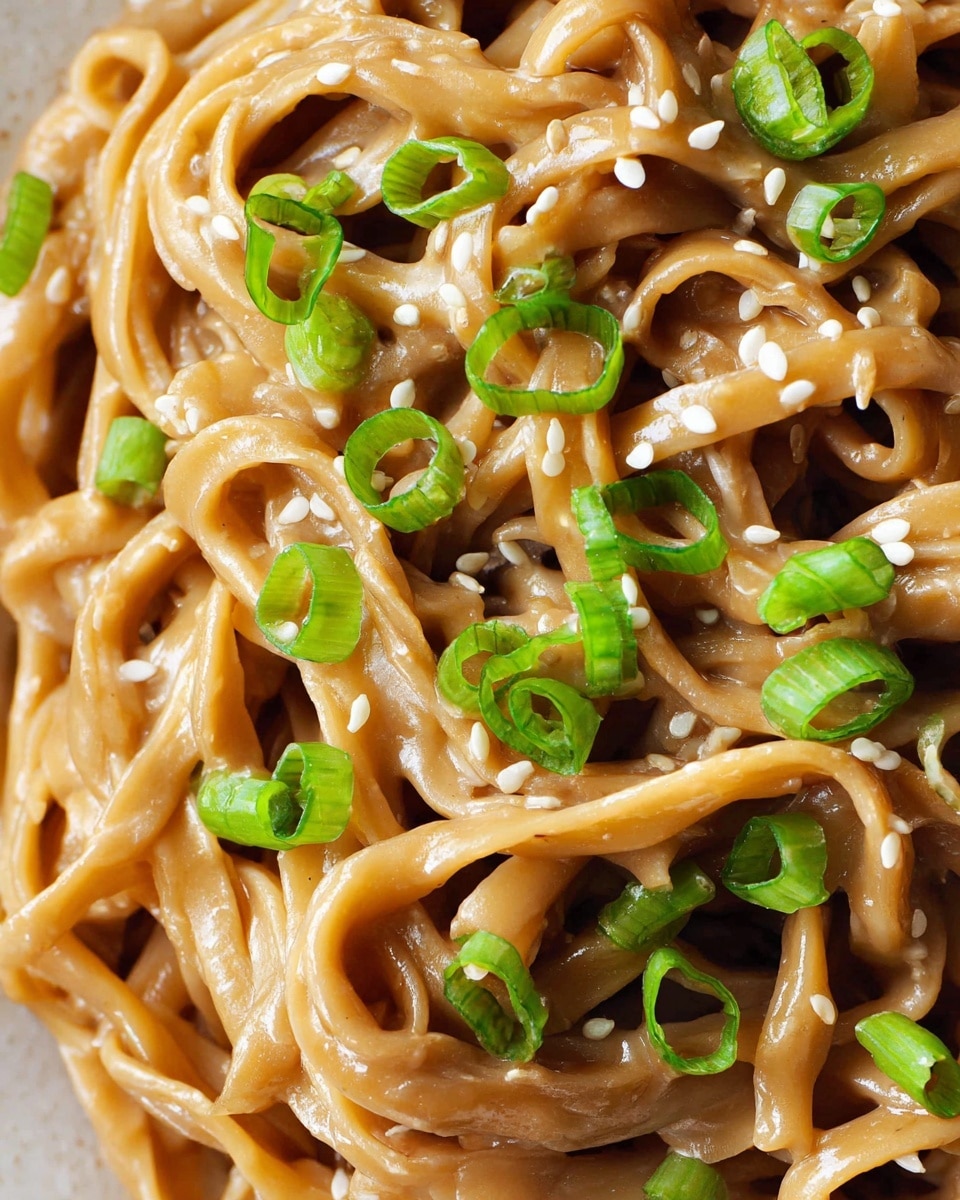 The image shows a close-up of thick noodles coated in a light brown creamy sauce. The noodles are tangled together in several layers, with a smooth and slightly glossy texture. On top, there are chopped bright green scallions scattered evenly, adding a fresh touch. Small white sesame seeds are sprinkled all over the noodles, creating a nice contrast. The background is a white marbled texture. Photo taken with an iphone --ar 4:5 --v 7