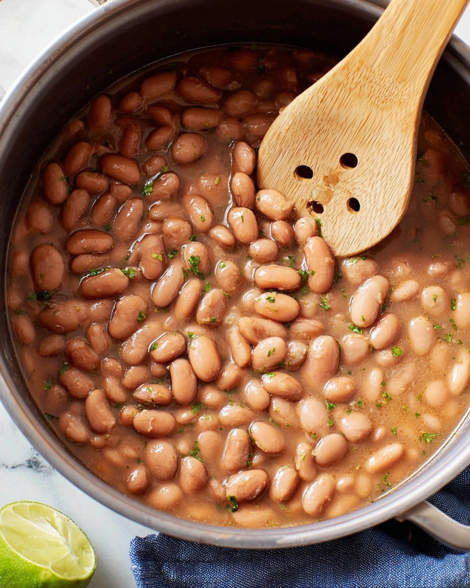 A close-up view of a pot filled with cooked pinto beans in a light brown, slightly thick broth, sprinkled with small pieces of green herbs scattered throughout. A light-colored wooden spoon with holes rests on top of the beans, partially submerged in the mixture. The pot sits on a white marbled surface with part of a sliced lime and a blue cloth visible near the edge. Photo taken with an iphone --ar 4:5 --v 7