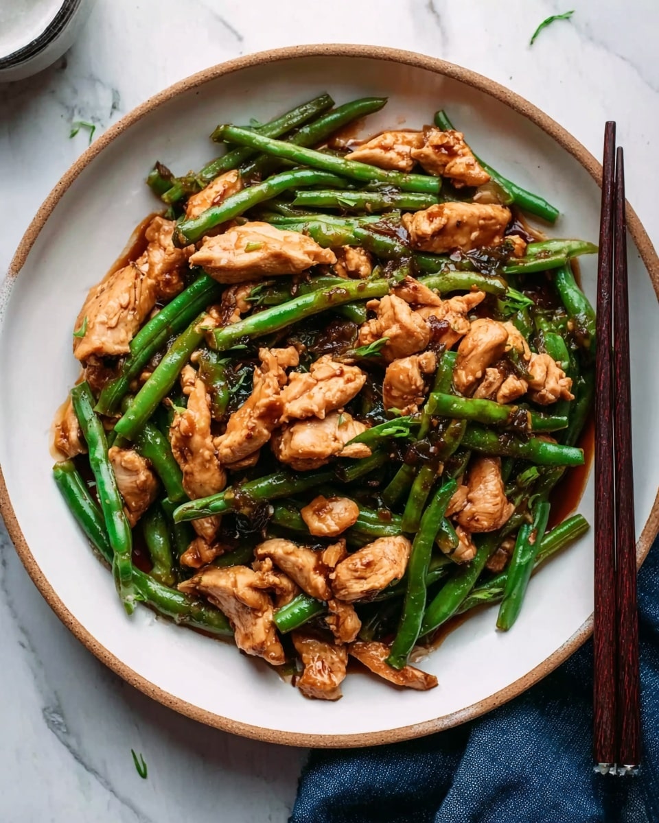 The image shows a white round plate filled with a stir-fry dish made of small pieces of cooked chicken and bright green beans. The chicken pieces are mixed evenly with the green beans and are coated in a shiny dark brown sauce, adding a rich color and texture. The chicken looks tender and slightly browned, while the green beans appear fresh and crisp. The plate is placed on a white marbled surface, with a pair of dark brown chopsticks resting on the right side of the plate and a small portion of a blue cloth visible in the corner. Photo taken with an iphone --ar 4:5 --v 7