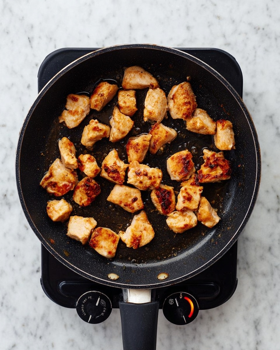 The image shows a black frying pan on a small electric stove with a dial set to a low number. Inside the pan are many small, browned pieces of cooked chicken spaced out on the surface. The chicken pieces have a golden-brown color with some darker grill marks and a slightly shiny texture from the cooking oil. The background is a white marbled surface. photo taken with an iphone --ar 4:5 --v 7