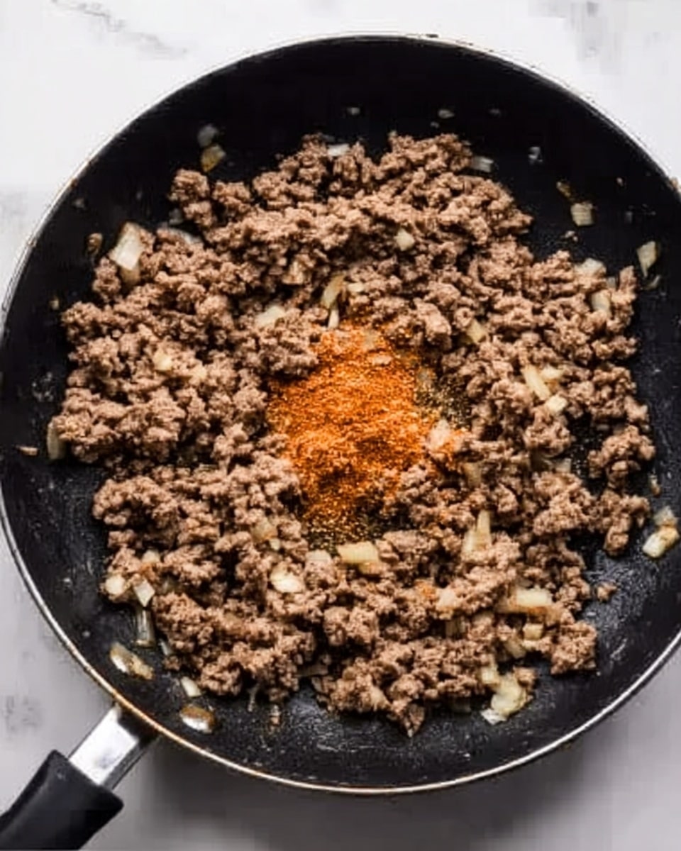 A close-up view of a black frying pan filled with cooked ground meat mixed with small bits of onions and spices scattered on top. The meat is brown with uneven texture, some parts look slightly more cooked than others. There is a small heap of orange-brown powder in the middle, likely seasoning, resting on the cooked meat. The frying pan sits on a white marbled surface, with no other items around. The handle of the frying pan extends to the left. photo taken with an iphone --ar 4:5 --v 7