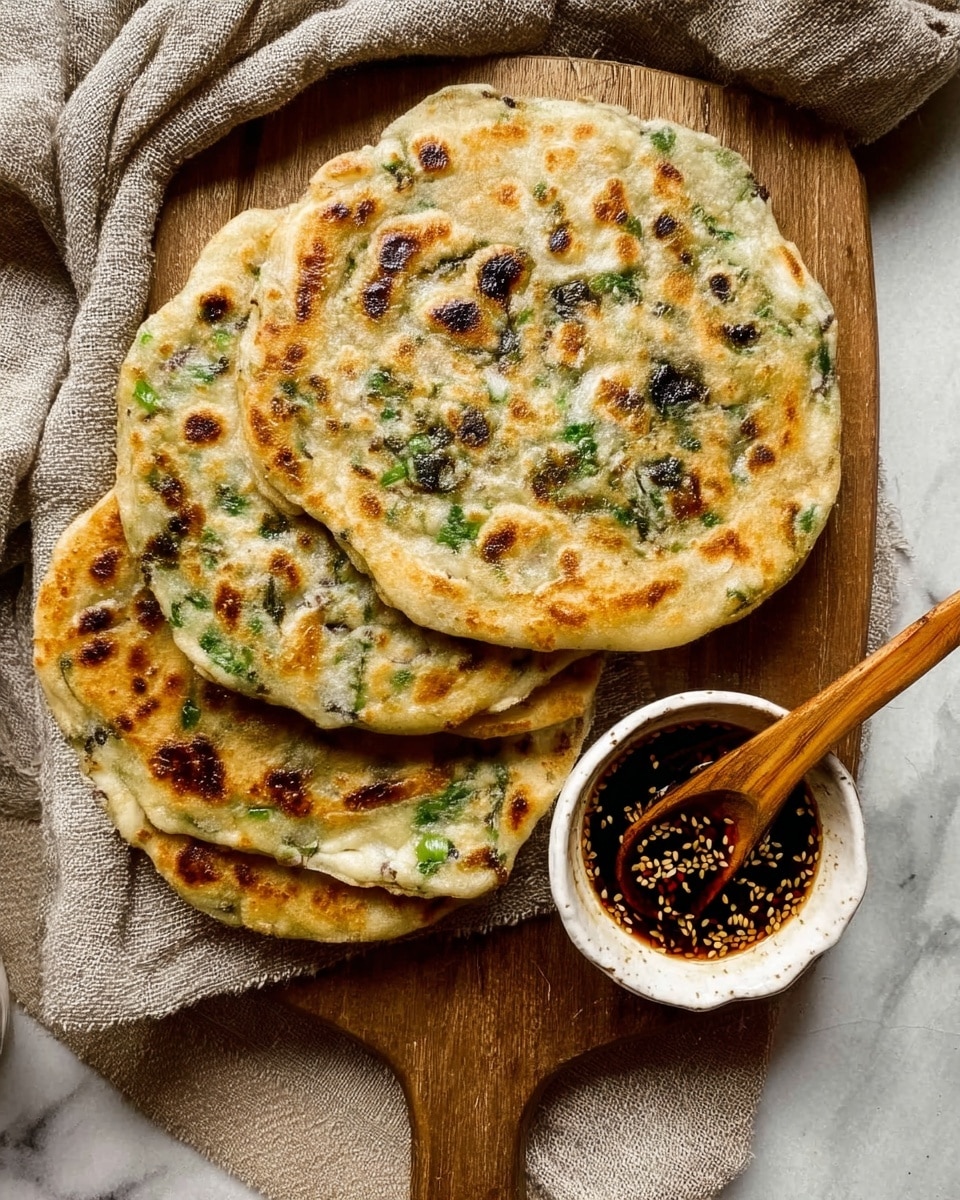 Three round flatbreads with golden brown spots and green pieces inside are stacked slightly on top of each other on a wooden board. Next to the flatbreads is a small white bowl filled with dark sauce that has sesame seeds on the surface, with a wooden spoon resting in the bowl. The board lies on a beige cloth, all placed on a white marbled surface. photo taken with an iphone --ar 4:5 --v 7