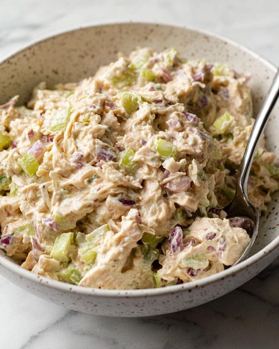 A close-up view of a white speckled bowl filled with a creamy, chunky salad that has a soft beige color with small pieces of light green celery and purple-red onion mixed throughout. The texture appears thick and slightly coarse, with visible shreds and bits of ingredients blending together. A metal spoon with a shiny surface is partially inserted in the salad on the right side of the bowl. The bowl is set on a white marbled surface. photo taken with an iphone --ar 4:5 --v 7
