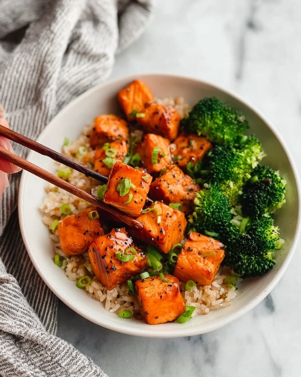 The image shows a white bowl filled with a bed of light brown rice at the bottom layer, topped with vibrant orange, grilled salmon cubes sprinkled with green onion slices, adding a fresh touch. To one side of the salmon and rice, there are bright green broccoli florets, adding a pop of color and texture contrast. A pair of chopsticks held by a woman's hand is lifting one salmon cube, showing its slightly glazed, seasoned surface with small black pepper flakes. The bowl is placed on a white marbled surface with a soft striped cloth nearby. Photo taken with an iphone --ar 4:5 --v 7
