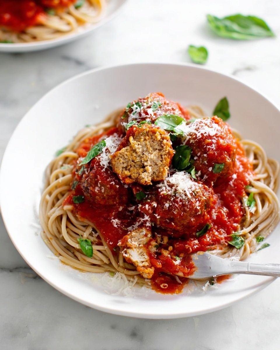 A white plate holds a nest of light brown spaghetti arranged in a circle at the bottom. On top, there are about five large meatballs covered in bright red tomato sauce with visible tomato chunks. One meatball is cut in half, showing a light brown, textured inside. The dish is sprinkled with white grated cheese and topped with fresh green basil leaves. A silver fork is placed on the right side of the plate, partly resting on the spaghetti and sauce. The background is a white marbled surface with another plate blurred in the background. photo taken with an iphone --ar 4:5 --v 7