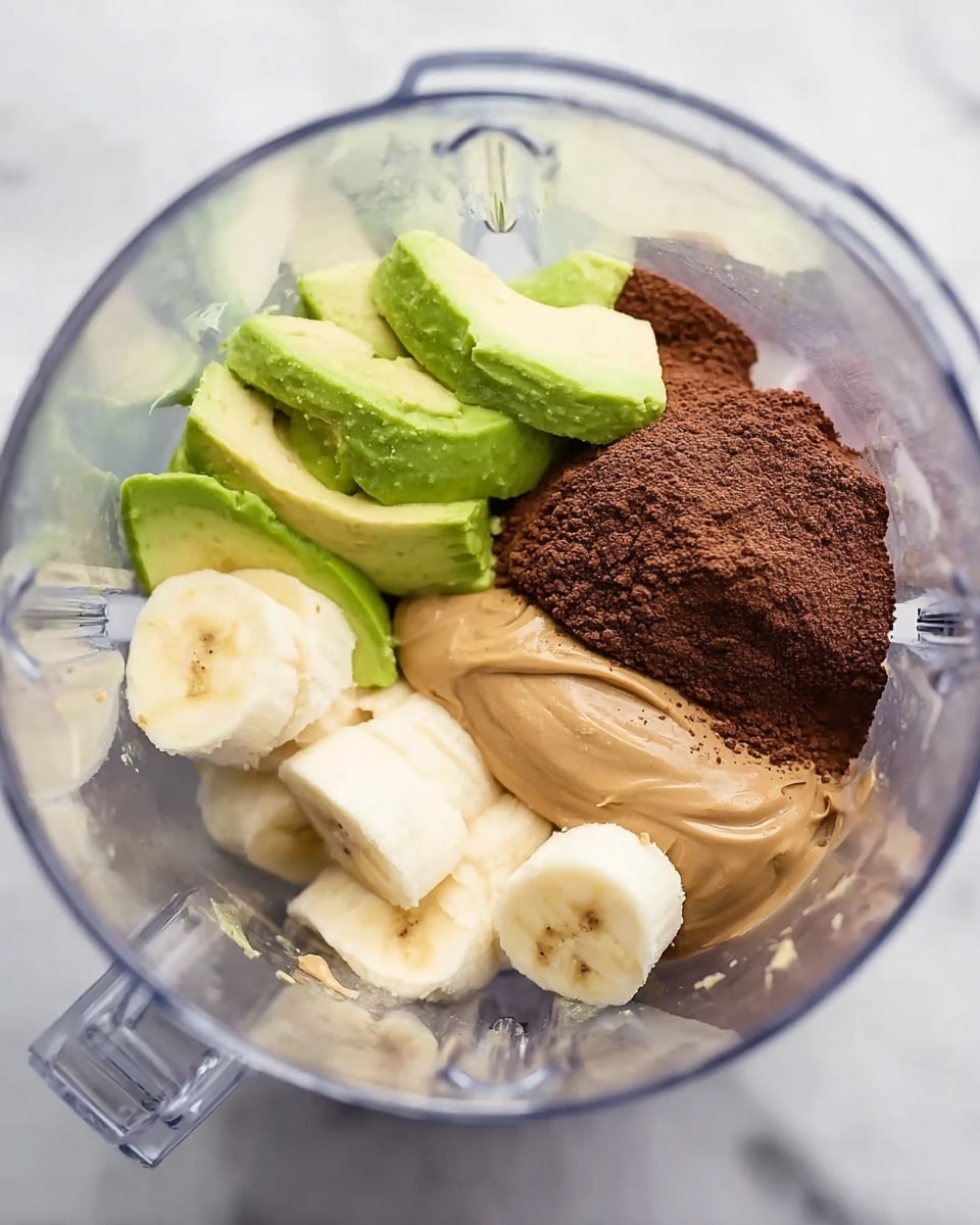 A clear blender container shows four main layers inside. On the bottom right, there are banana slices in pale yellow with soft textures. Above the bananas, on the right side, is a smooth, creamy layer of light brown peanut butter. To the left of the bananas, there is a dark brown powdery layer of cocoa. The top left side contains slices of green avocado with smooth, creamy texture and lighter green inner flesh. The blender is placed on a white marbled surface. photo taken with an iphone --ar 4:5 --v 7
