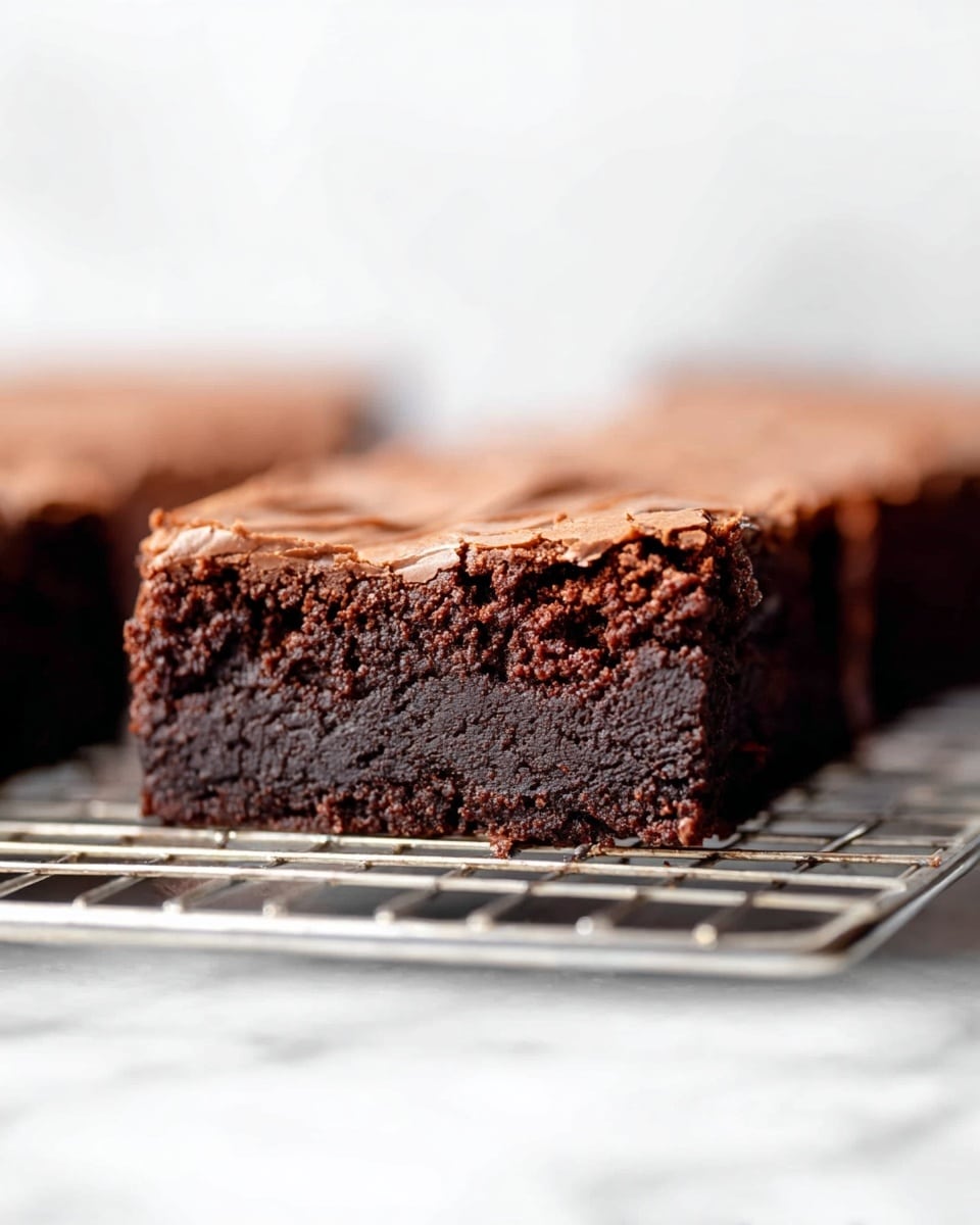 A close-up of a square slice of rich brownie resting on a silver cooling rack, placed over a white marbled surface. The brownie has two layers: the bottom layer is dark brown with a moist, slightly crumbly texture, showing tiny cracks and a dense look, while the top layer is a lighter brown with a smooth, creamy, and slightly wavy surface. The background is softly blurred with more brownie pieces visible, emphasizing the main brownie slice in sharp focus. photo taken with an iphone --ar 4:5 --v 7