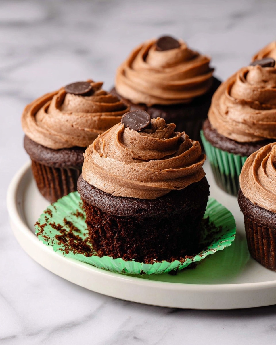 The image shows several chocolate cupcakes with a dark brown, moist cake base, each topped with a thick swirl of smooth, light brown chocolate frosting. One cupcake in the foreground has its green wrapper partly peeled back, revealing the rich cake inside. The cupcakes sit on a white ceramic plate against a white marbled surface. The frosting is textured with swirls and slight peaks, and the cupcake in front has two small chocolate pieces on top. Photo taken with an iphone --ar 4:5 --v 7