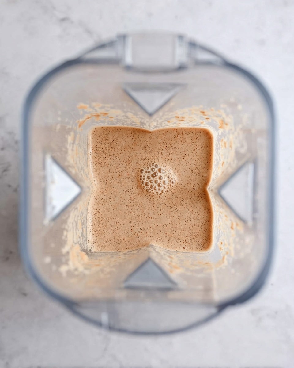 A clear blender jar seen from above contains a light brown, slightly bubbly mixture that sits mostly flat inside, with small chunks stuck around the sides of the jar. The jar has a square base visible through the mixture and is placed on a white marbled surface. The overall texture of the mixture looks smooth but with some small lumps spread evenly throughout. photo taken with an iphone --ar 4:5 --v 7