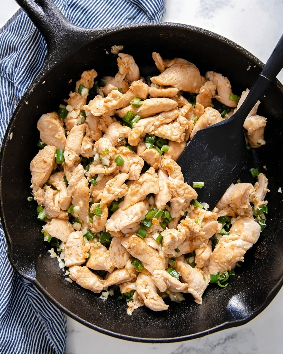 The image shows a black cast iron pan filled with cooked light brown sliced chicken pieces mixed with small chopped green onions and bits of sautéed garlic. The chicken slices have a soft texture with a slight golden edge, layered loosely inside the pan. There is a black spatula lifting some chicken near the top right, the pan is placed on a white marbled surface with a blue and white striped cloth partially visible in the upper left corner. Photo taken with an iphone --ar 4:5 --v 7