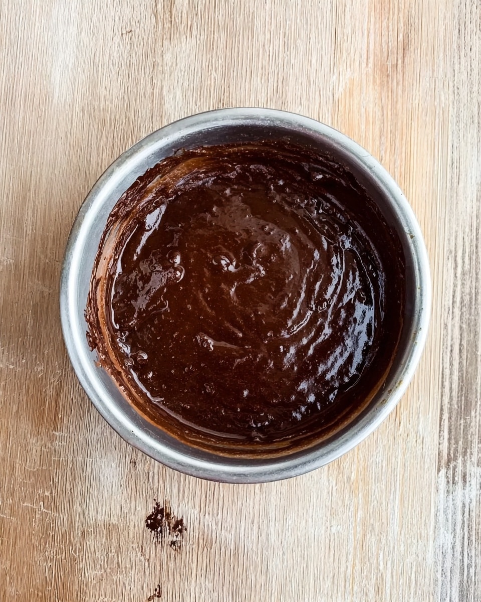 A round silver bowl is shown from above filled with thick, dark brown chocolate batter that has a smooth yet slightly uneven texture on the surface. The bowl rests on a wooden surface with light, natural wood grain. There is a small drip of chocolate batter on the inside rim and side of the bowl, adding an organic touch. The overall look is rich and creamy, with shiny highlights on the dark chocolate mix. Photo taken with an iphone --ar 4:5 --v 7