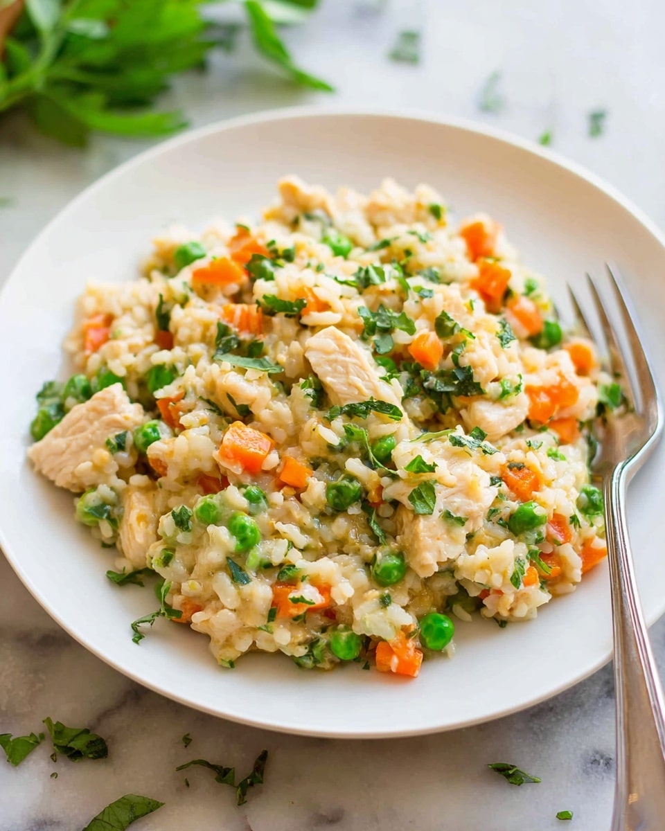 A close-up of a white plate filled with creamy rice mixed with small pieces of light beige chicken, bright orange carrot chunks, and vibrant green peas, all lightly sprinkled with finely chopped green herbs. The dish has a soft, moist texture with a mix of colors from the vegetables and meat spread evenly throughout the rice. A silver fork rests on the right side of the plate. The plate is placed on a white marbled textured surface with a few green herb leaves slightly out of focus in the background. photo taken with an iphone --ar 4:5 --v 7