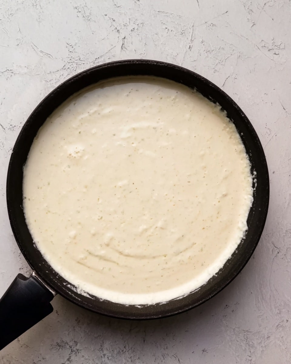 A black skillet with a thick, creamy white sauce covering the entire inside surface, showing slight uneven texture and small bubbles, placed on a white marbled surface. The sauce has a smooth top with some light cream-colored specks throughout. Photo taken with an iphone --ar 4:5 --v 7